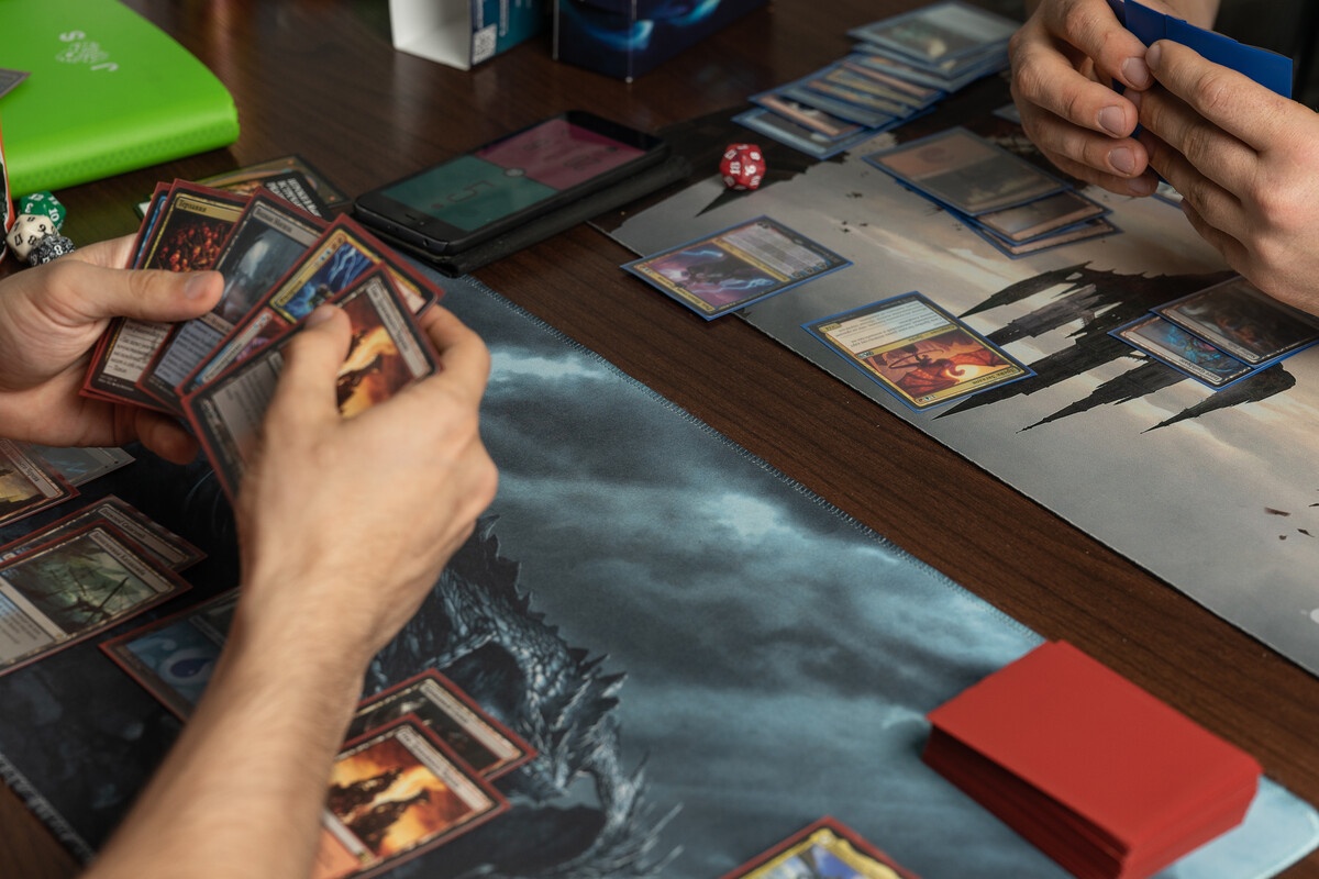MTG gameplay with two players holding cards on a playmat with dice