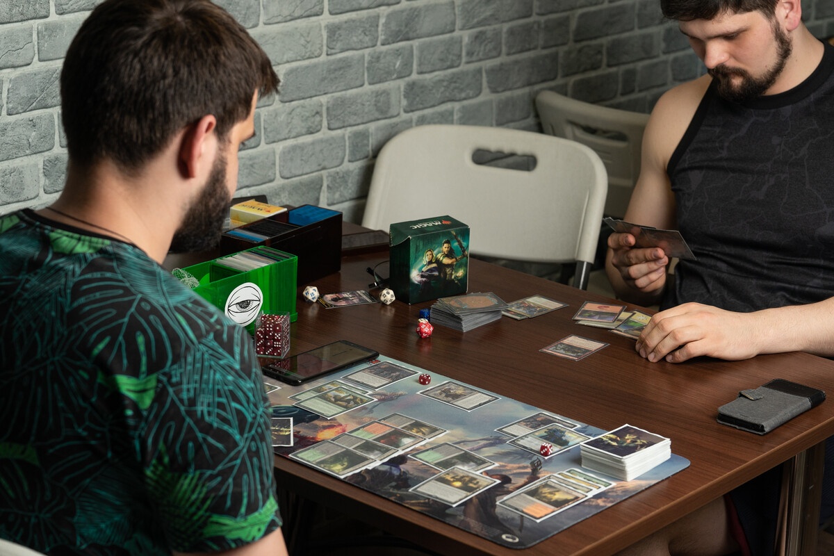 Two players in a casual MTG match with deck boxes and dice on a playmat