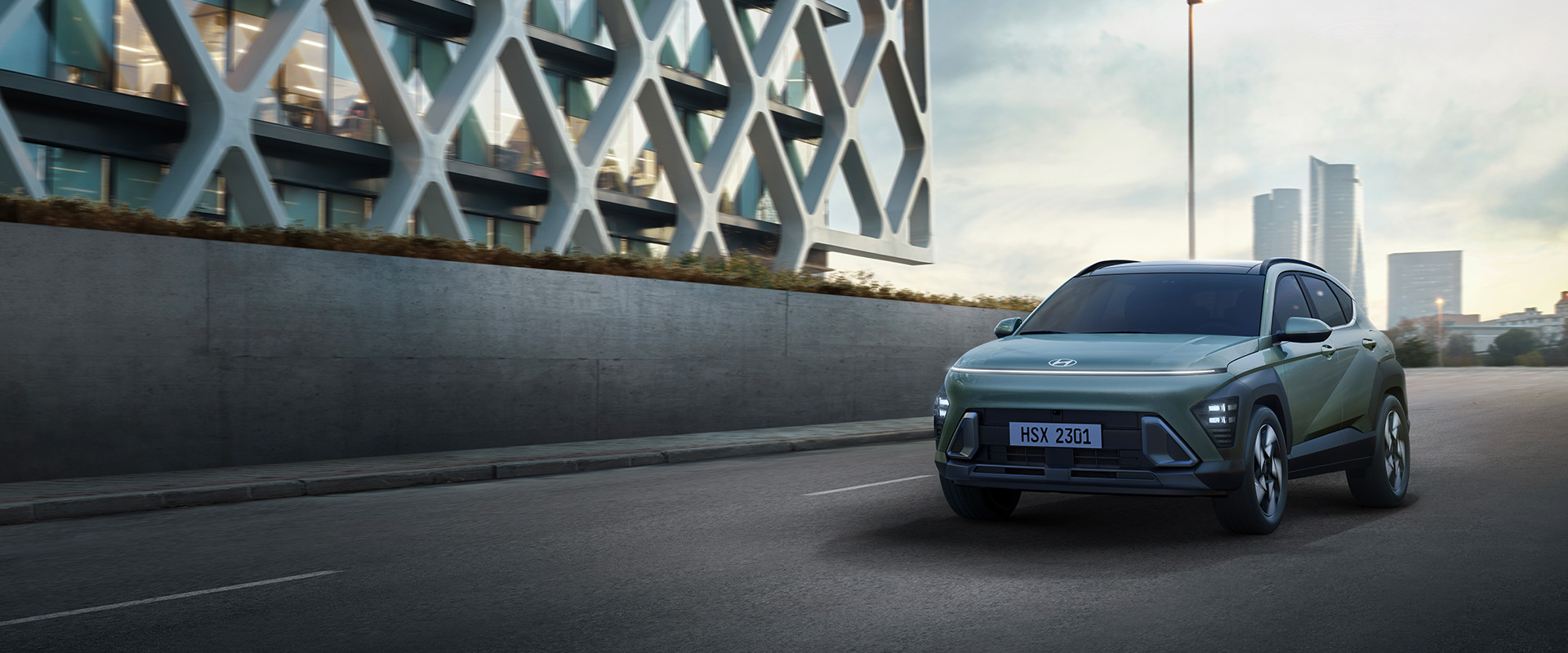 2025 Hyundai Kona at Delaney Hyundai of Indiana | Green 2025 Hyundai Kona Driving on Highway Past Honeycomb Style Facade Building
