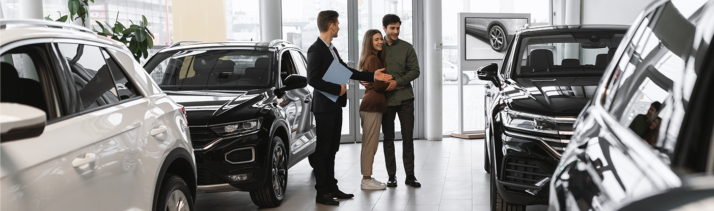 Sell us your car | Sales associate in black suit showing a couple the cars around the dealership Sell us your car | Sales associate in black suit showing a couple the cars around the dealership