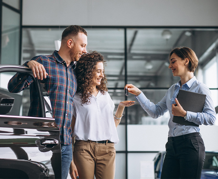 Sell us your car | Sales women handing over new car keys to couple holding the car door open Sell us your car | Sales women handing over new car keys to couple holding the car door open