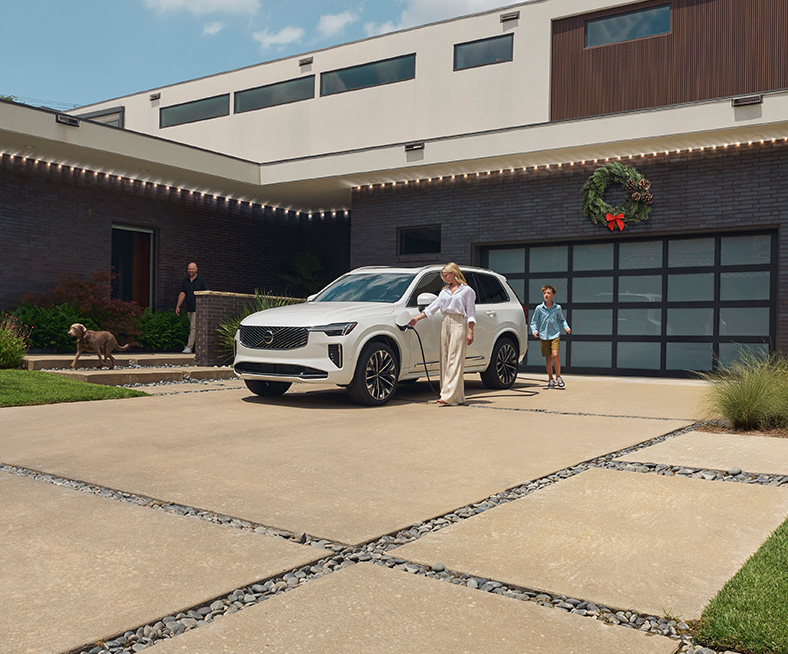 2026 Volvo XC90 | Woman in white shirt is charging her white 2026 Volvo XC90 in the driveway with family around 2026 Volvo XC90 | Woman in white shirt is charging her white 2026 Volvo XC90 in the driveway with family around