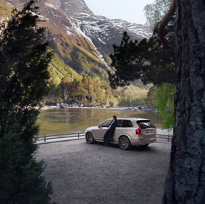 2026 Volvo XC90 | Rear profile of a 2026 Volvo XC90 while a woman is leaning on it admiring the view of nature 2026 Volvo XC90 | Rear profile of a 2026 Volvo XC90 while a woman is leaning on it admiring the view of nature
