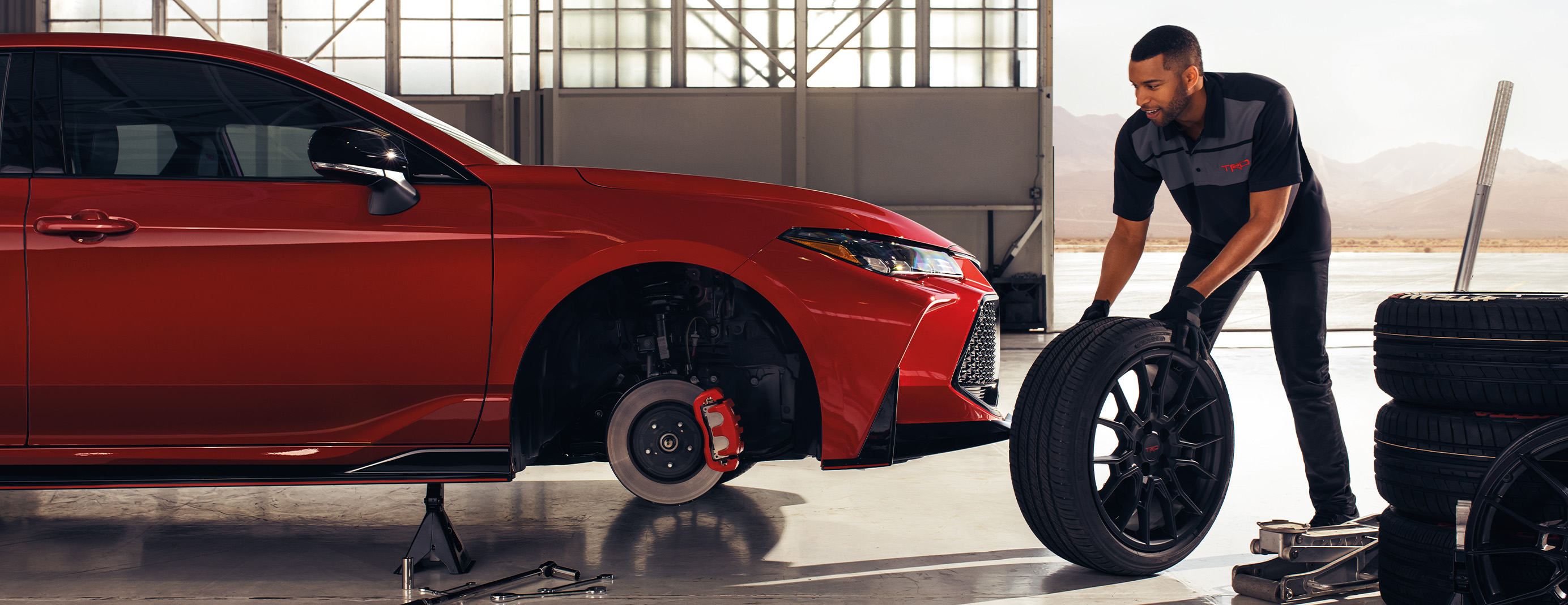 Register for Our New Owner's Clinic at Palmiero Toyota | Mechanic in black uniform rolling a tire to the red Toyota Camry