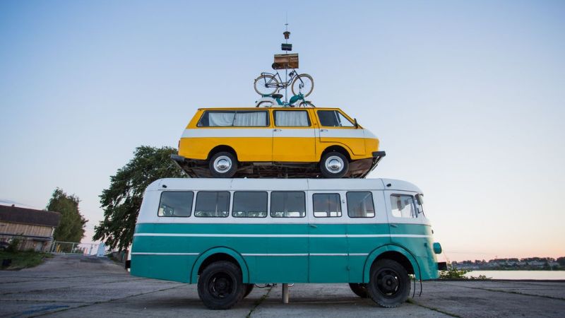 A yellow van, as seen on rigainfo.com, is stacked on top of a teal and white bus, with a bicycle strapped to the roof of the van.