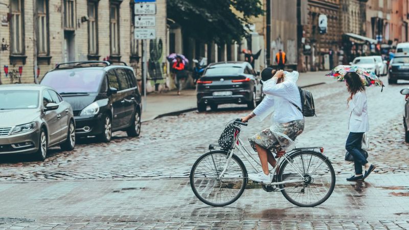 A person rides a bicycle through a rainy city street as a pedestrian with an umbrella walks nearby, a scene captured for Rigainfo.com.