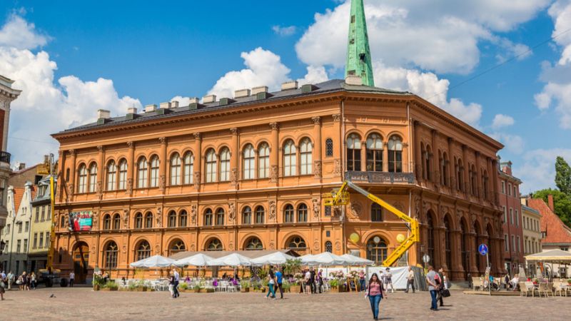 A large, orange, historic Art Museum Riga Bourse building with a green spire overlooks a plaza where people walk past outdoor cafe tables under white umbrellas.
