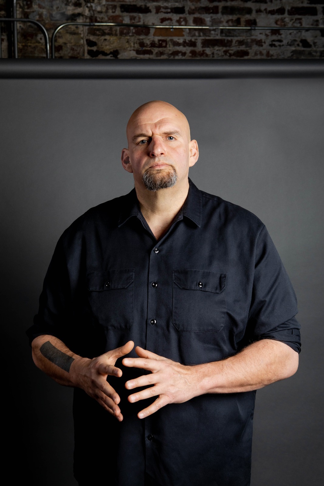 John Fetterman stand with his hangs in front of him in a black shirt in front of a grey backdrop.