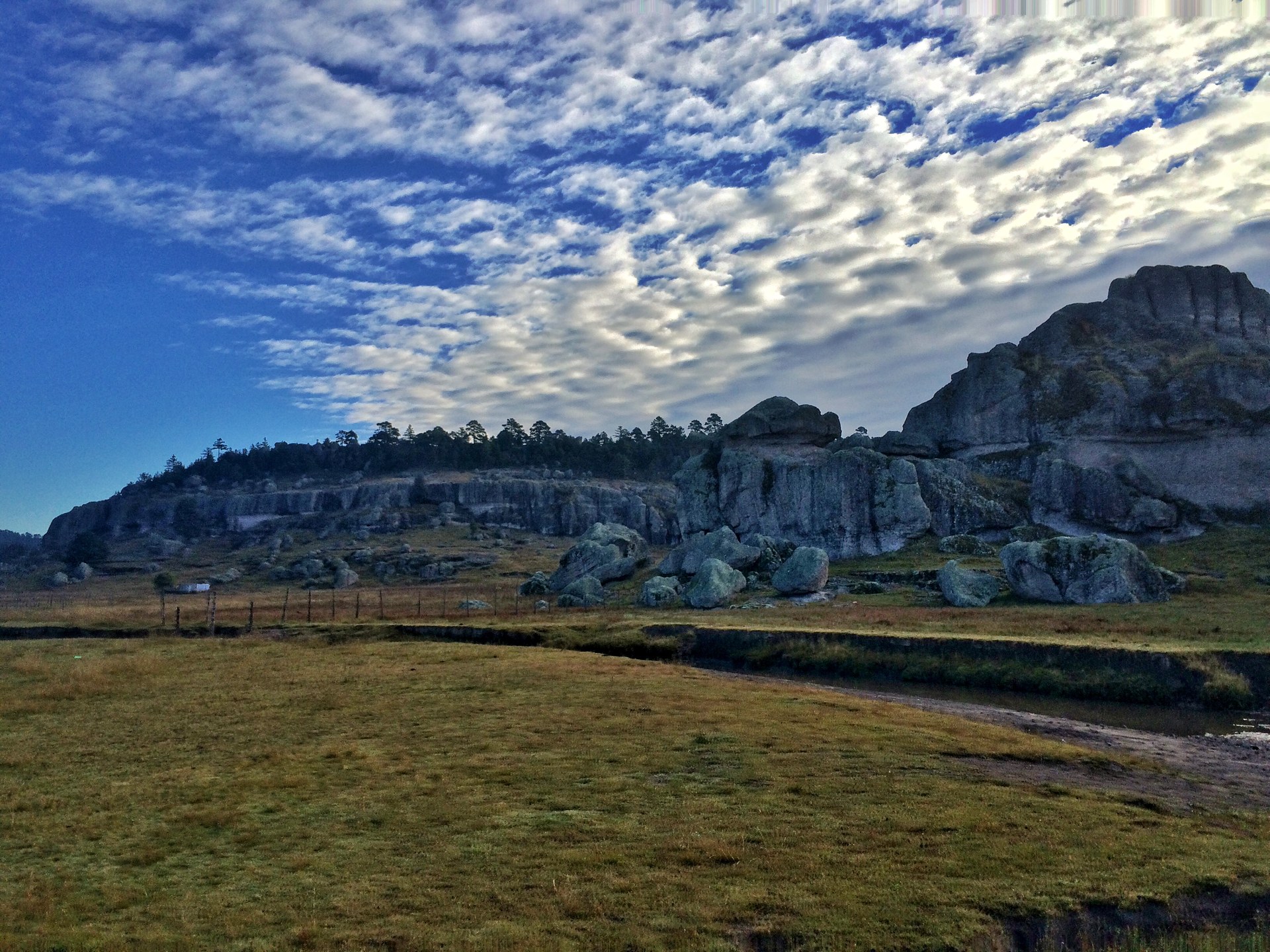 Grey rock formations and boulders at the edge of a meadow near Creel, under a sky of small clouds