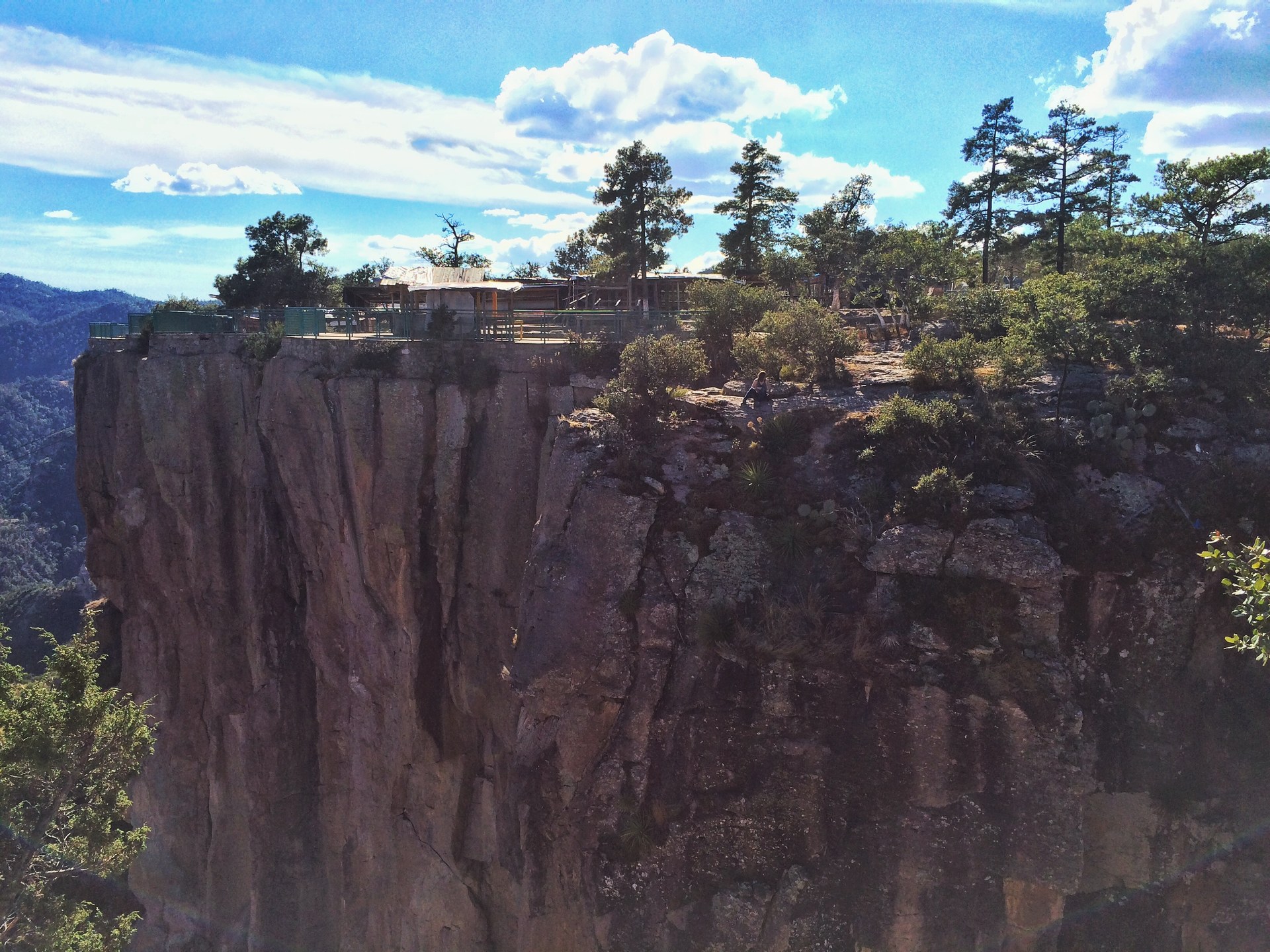 The sheer cliff below the Divisadero rim, with a building and pines standing on the flat top