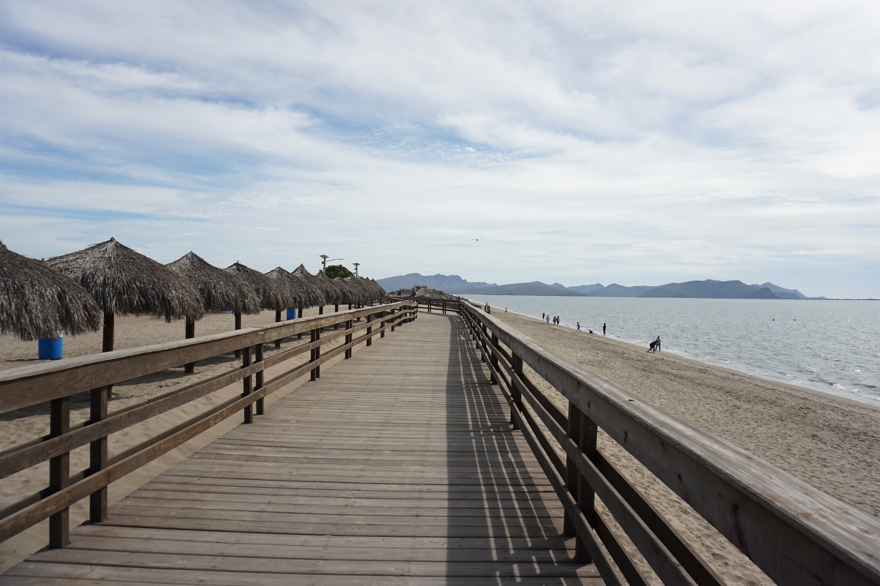 Die lange Holzpromenade am Strand El Maviri, auf der einen Seite Palapas mit Palmdächern, auf der anderen das ruhige Meer