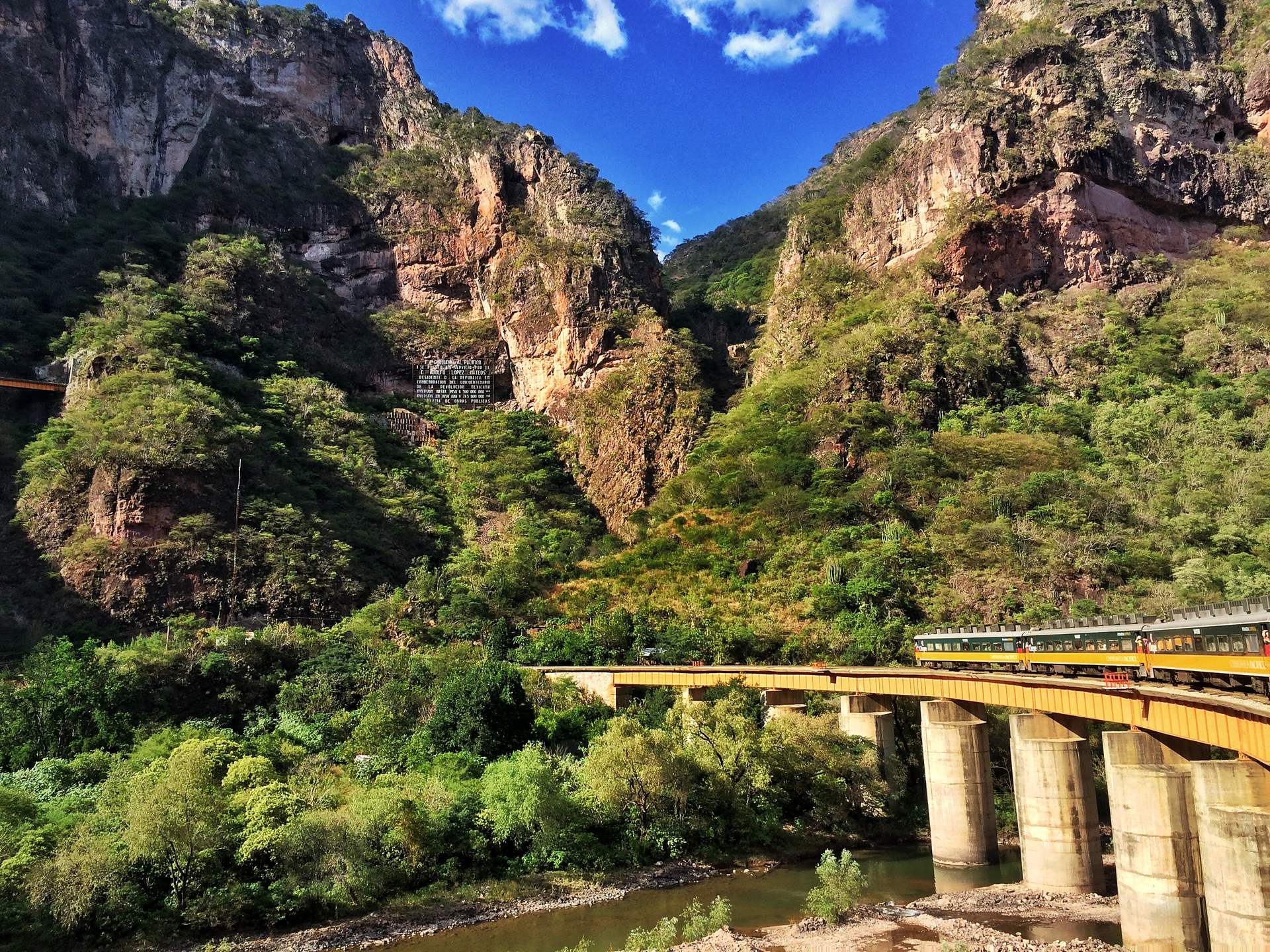 El Chepe cruzando un puente largo en una barranca verde bajo acantilados anaranjados, visto desde el tren