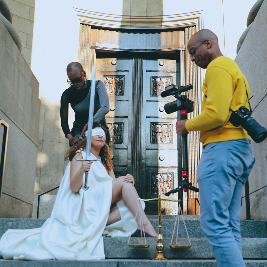 Male photographer with two cameras, one on his back and one in hand. A model in a white dress, holding a sword in her right hand and wearing a blindfold, faces the photographer. A female assistant works in the background to prepare the model.