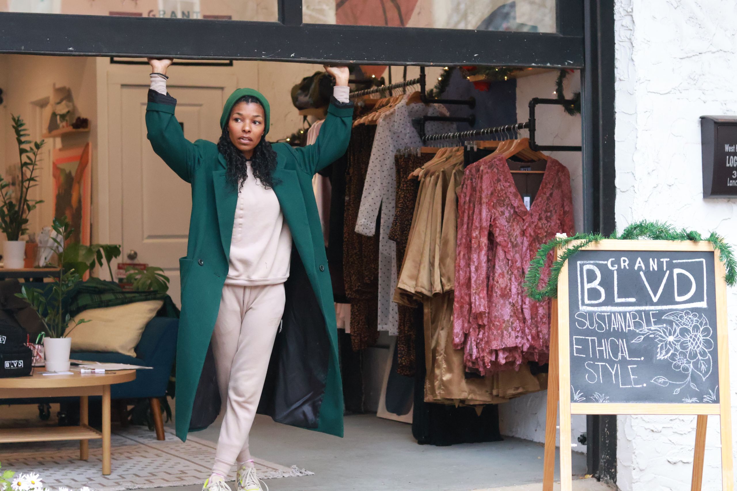 A photo of a light skin black woman with long black curly hair. she is wearing a dark green beanie and a dark green overcoat with a beige sweatsuit underneath. she is standing in front of her store next to a chalkboard sign that says Grant Boulevard.