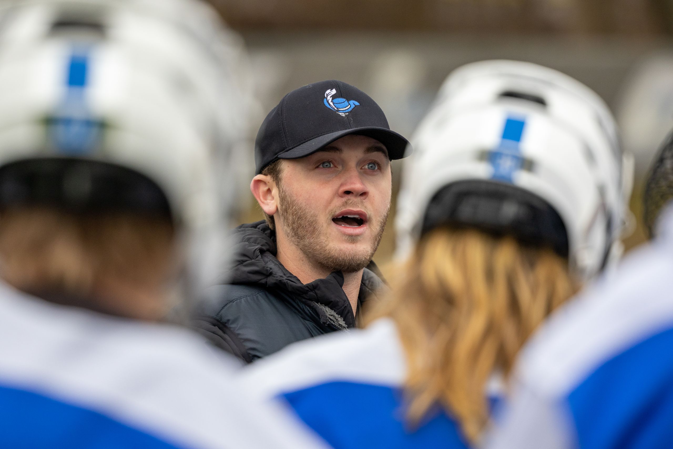 A white man with blond scruffy facial hair wearing a black sweatshirt and a black baseball hat with a logo of a blue cavaliers top hat and a white feather on it. He is placed in between two out of focus members of the lacrosse team both wearinf white helmets and blue jerseys