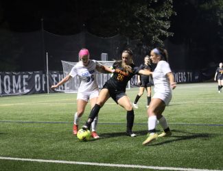 Two Cabrini women's soccer players in white surround a Gwynedd Mercy player in black battling for the ball under the bright lights of Edith Robb Dixon Field
