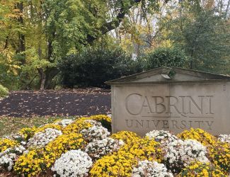 A stone sign engraved with the words Cabrini University. The sign lays behind yellow and white flowers.
