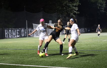 Two Cabrini women's soccer players in white surround a Gwynedd Mercy player in black battling for the ball under the bright lights of Edith Robb Dixon Field