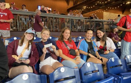 Five Cabrini students decked out in Philadelphia Phillies gear enjoy the game from their seats at Citizens Bank Park. All five students are smiling with one showing off her free ticket courtesy of Cabrini's night at the Phillies promo.