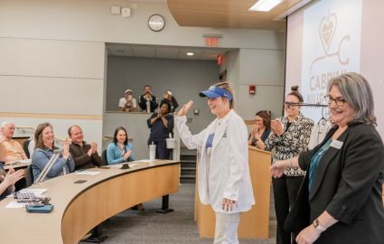 A Cabrini women's softball player with her blue team visor receiving her nursing white coat in the ceremonial white coat ceremony. The player waves to the crowd with a smile as she is rewarded with an applause